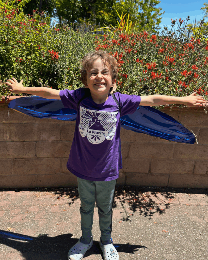 young white boy is posing with dragon fly wings on, arms outstretched, and a huge smile