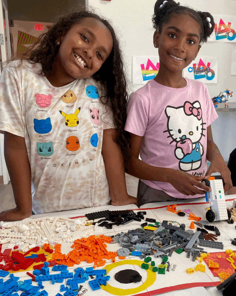 two young black girls smile behind a table with colorful lego pieces spread across it