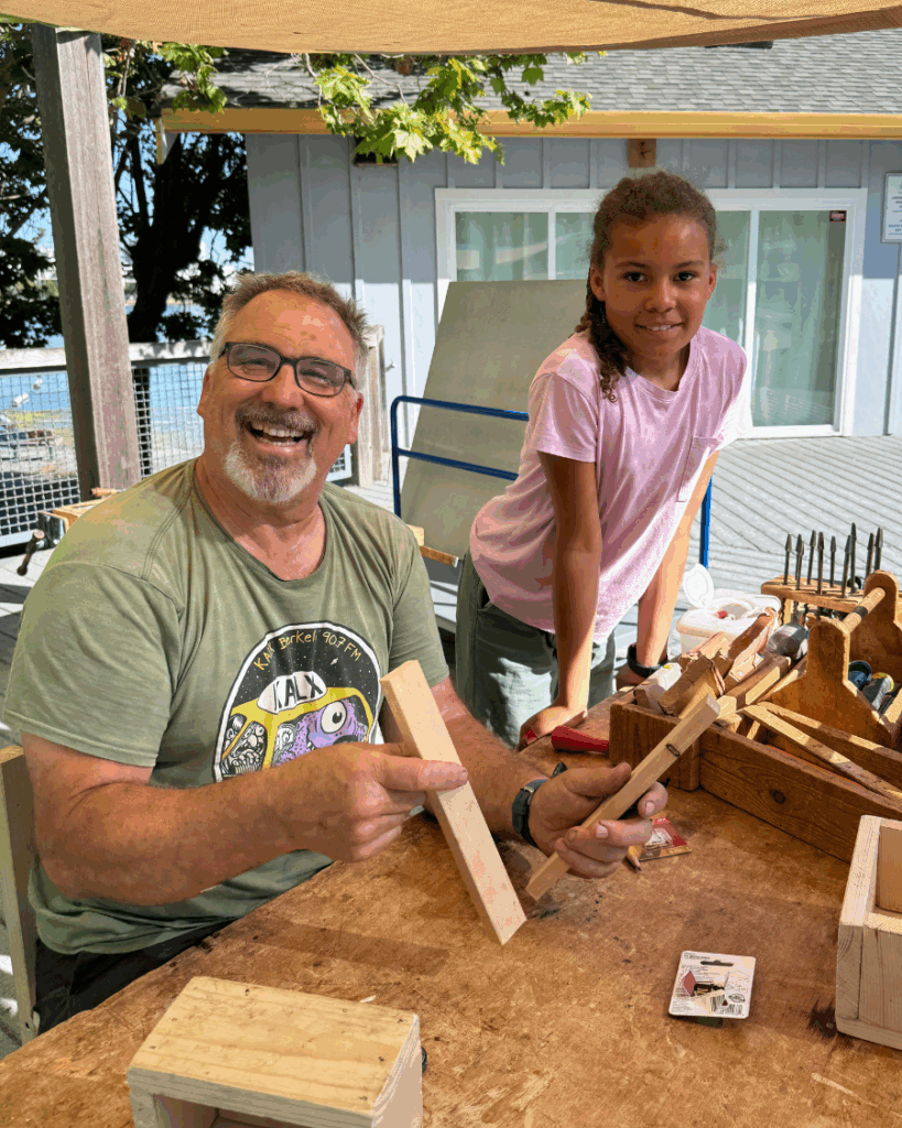 Carpentry teacher and student smile at the camera in front of various carpentry tools