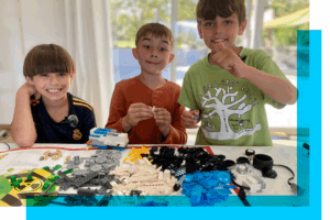 photo of three young white boys behind a table filled with legos giving thumbs up and smiling