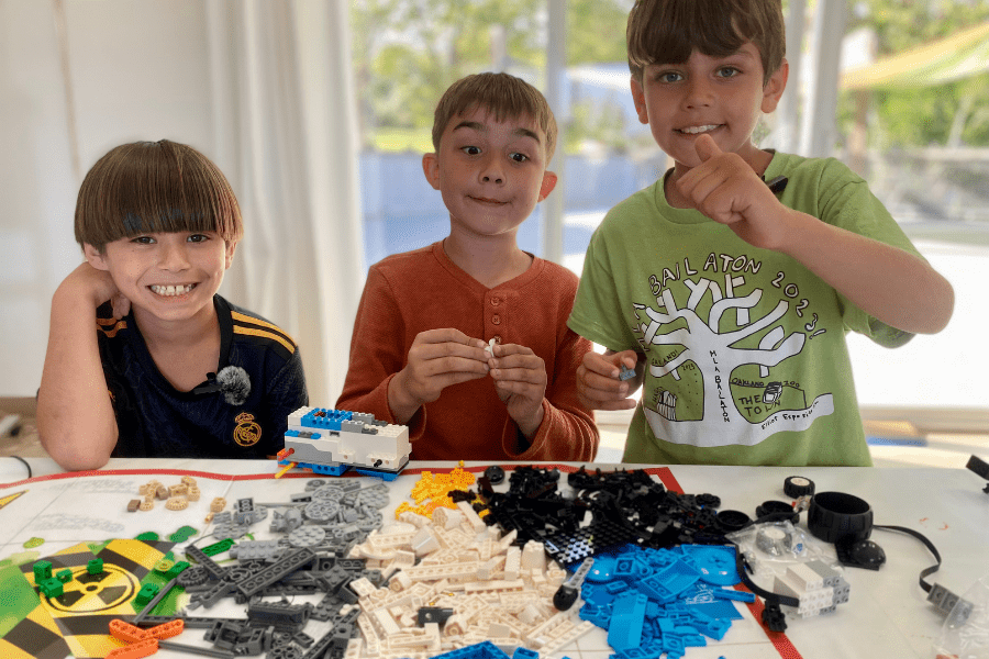 website 600x900 three young white boys behind a table filled with legos smiling and giving thumbs up!